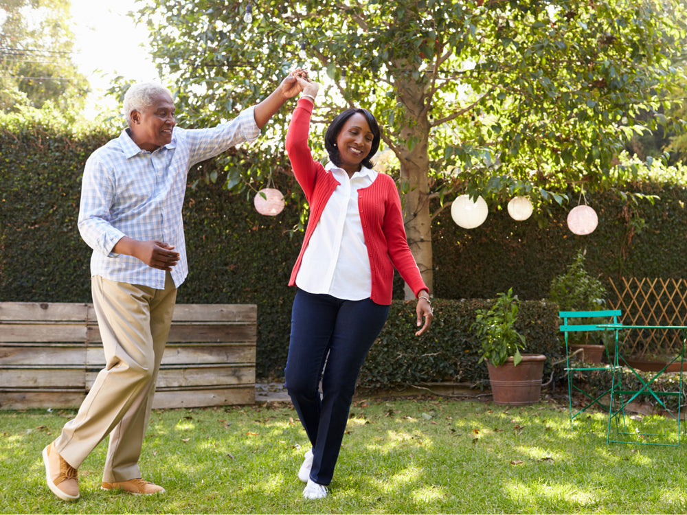 Senior couple dancing in a garden with greenery and a wooden bench in the background.