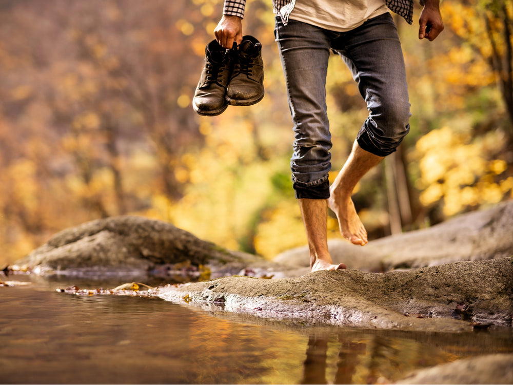 Person walking barefoot over water with shoes in hand, autumn foliage in the background