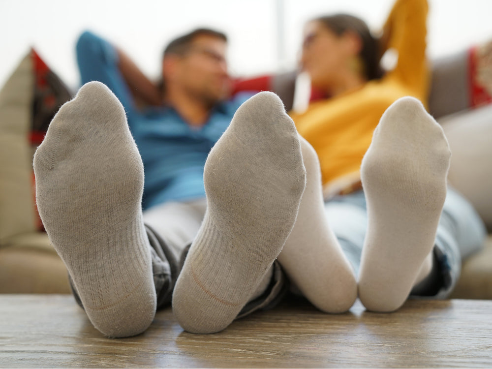 Two pairs of feet wearing socks on a wooden floor with people sitting in the background.